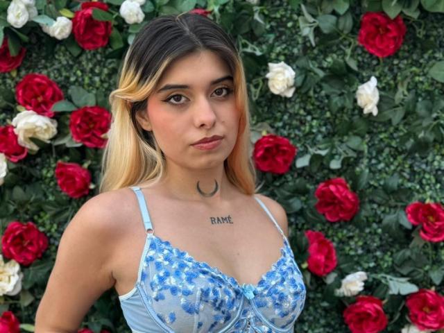 LannaHamilton stands in front of a wall of red and white roses, wearing a light blue lace bra with blue floral patterns. She has long, blonde hair with dark roots and a small crescent moon tattoo on her chest, just below her cleavage. Her brown eyes are slightly downcast, and she has a neutral expression. The image is sharply focused on her upper body, highlighting the detailed lacework of her bra and the natural greenery of the rose-filled background. The composition uses contrasting colors to draw attention to her and the floral wall.