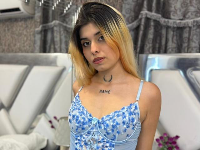 LannaHamilton stands in a room with a silver-patterned curtain. She has long, blonde-to-black ombre hair and a light skin tone. She wears a light blue, floral-patterned bra with lace detailing. A small crescent moon tattoo is centered on her upper chest, with "BAME" tattooed above it. Her expression is neutral. The background includes a silver headboard and a white air conditioner. The composition focuses on her upper body.