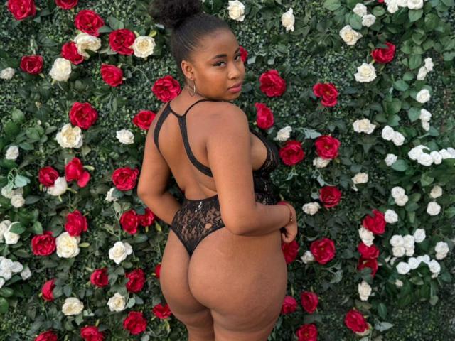 RailyBenet has dark skin, a curly bun, and wears a black lace thong with a cutout back, showing her big ass and small thong. Red and white roses surround her.