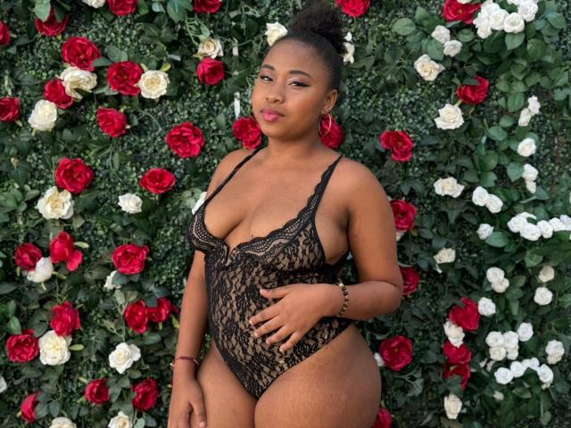 RailyBenet with dark skin, big ass, and small tits in a black lace one-piece, standing against a wall of red and white roses, looking sexy with a bun.