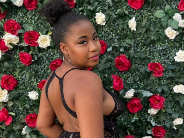 RailyBenet with dark skin, curly hair in a bun, wearing a black lace dress, showing her back with a side glance, red and white roses in the background.