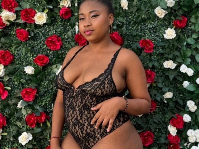 RailyBenet with dark brown skin, wearing a black lace one-piece, showing her boobs and a slight belly, standing against a red and white rose wall. She's looking confident.