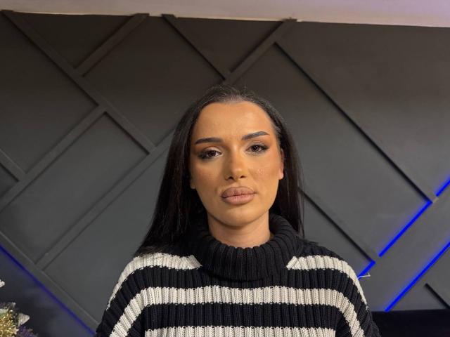 NaughtyyChloe, com cabelos longos e pretos, vestindo um suéter de malha listrado em preto e branco, tem lábios carnudos e olhos castanhos, e está em frente a uma parede escura com um padrão geométrico..
