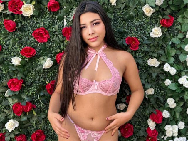 AmarantaRuiz, com cabelos longos e escuros, usando um sutiã e calcinha de renda rosa, mostrando seus seios e vulva, está em pé em frente a rosas vermelhas e brancas, com as mãos nos quadris, sorrindo..