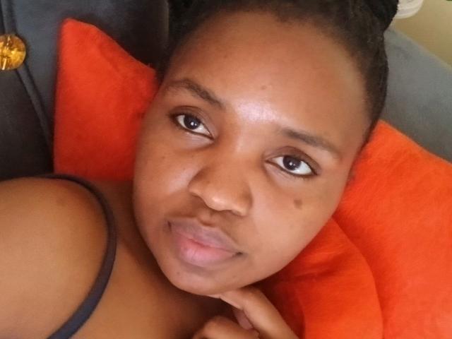 Close-up of LadyValour, a young woman with dark brown skin, brown eyes, and small, slightly parted lips. She has small, dark brown eyebrows and a small mole on her right cheek. Her hair is in a neat, small bun. She's lying on a bright orange pillow with her head resting on her right hand. She's wearing a dark strap, possibly from a tank top. The background is mostly out of focus, with a dark gray surface and a small, gold object in the top left. The composition is centered on her face, with a soft, natural light highlighting her features.