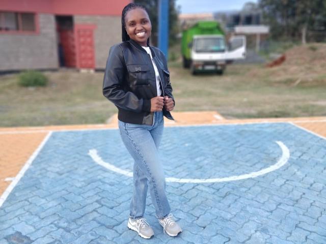 LadyValour stands on a blue-tiled court, wearing a black leather jacket over a white shirt, blue jeans, and grey sneakers. She has dark skin, a bright smile, and her hair is pulled back. The background includes a red building, green truck, and blurred greenery. Her relaxed pose with hands near her jacket adds a casual vibe. The court's blue tiles have a white circular design.