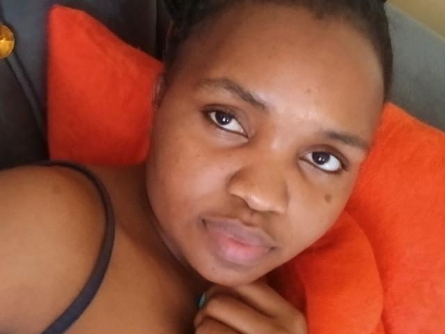 Close-up of LadyValour, a Black woman with dark brown skin and a small beauty mark on her right cheek. She has dark, slightly curly hair pulled back, and is looking to the right with a neutral expression. She's wearing a black spaghetti strap, resting her chin on her right hand. She's lying on an orange pillow, which is visible behind her head. The lighting is even, highlighting her smooth skin and natural makeup. The composition is centered, focusing on her face. The texture of the orange pillow is slightly fuzzy.