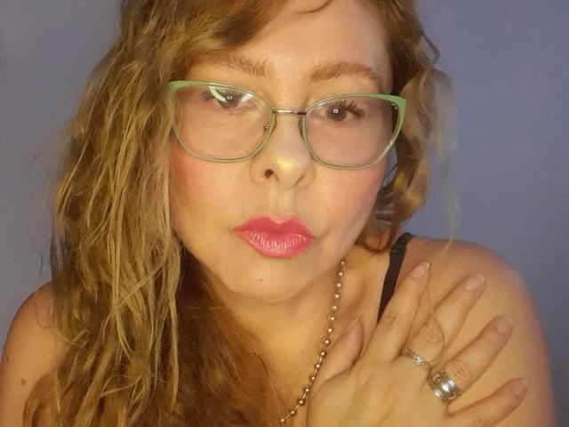MiaAmoretti with curly brown hair, green glasses, pink lipstick, pouty expression, black tank top, silver necklace, and ring, holding her shoulder against a plain background.