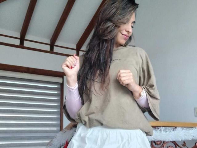 AntoniaHoney stands in a room with wooden beams and a slatted window. She has long, wavy brown hair and a light skin tone. She wears a loose, beige t-shirt over a pink long-sleeve top and white pants. Her hands are raised, fists slightly clenched, and she smiles softly. A gold bracelet is on her right wrist. The room has a white ceiling and a wooden railing. The composition focuses on AntoniaHoney, with natural light highlighting her relaxed, casual style.