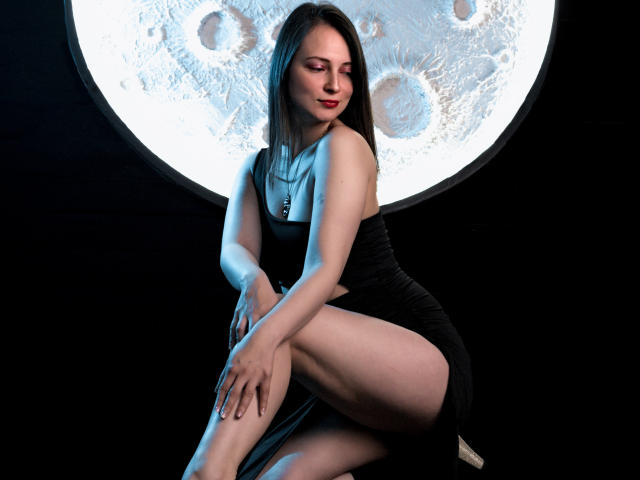 AmbeerWood with pale skin, black dress, and red lipstick, sitting with legs crossed, showing her ass, in front of a large moon-like light, looking down with a slight smile.