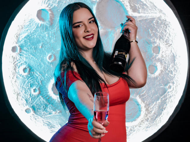 AuroraSantorini, com longos cabelos pretos, vestido vermelho, segurando uma garrafa na mão direita e oferecendo um copo na mão esquerda, sorrindo, com um fundo de lua, iluminação azul e branca, exibindo seus seios e nádegas..