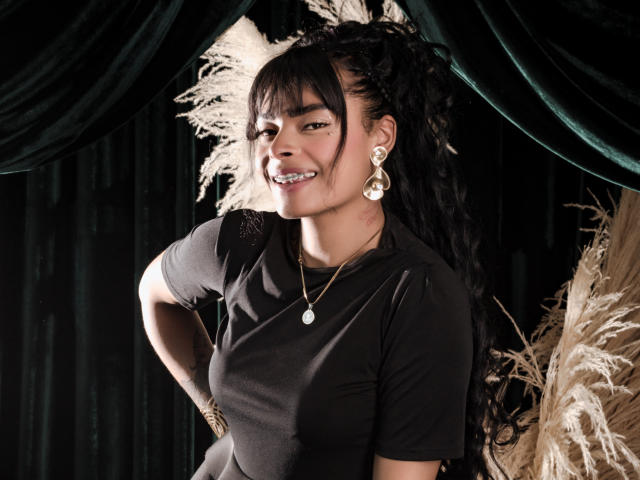 EmmaEbony with dark skin, black hair in a high pony, smiling, wearing a black shirt, gold earrings, necklace, and gold bracelet, standing against dark green curtains.