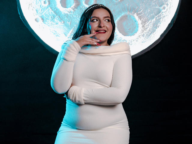 VictoriaRiver with tits in a white ribbed dress, touching her red-lipped mouth, smiling, black hair, white headband, moon-like light behind her, standing against dark background.