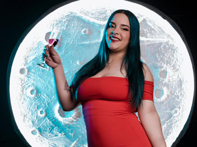 AuroraSantorini, con cabello azul turquesa, vestido rojo con un hombro descubierto, sosteniendo una copa de vino rosado, sonriendo, con los senos visibles, tatuaje en el brazo derecho, de pie frente a una luz circular..