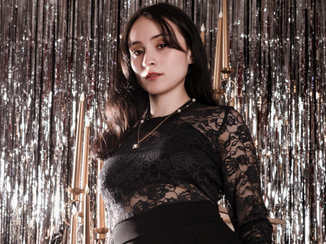 AnnieLamberth stands in front of a metallic, reflective curtain. She has long, dark hair and a fair skin tone. She wears a black lace top with floral patterns, a black skirt, and a gold necklace with a small pendant. Her expression is neutral, with slightly parted lips. The lighting highlights her face and the lace texture. The image is sharp with a focus on AnnieLamberth and her detailed lace top. Her posture is relaxed, with her right hand behind her. The metallic curtain adds a shiny, reflective background.