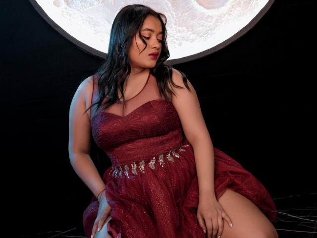Sarita, une femme séduisante avec des courbes, portant une robe rouge en dentelle, assise, regardant vers le bas, cheveux noirs ondulés, fard à paupières rouge, poitrine généreuse, cuisse visible, ceinture argentée, fond évoquant la lune..