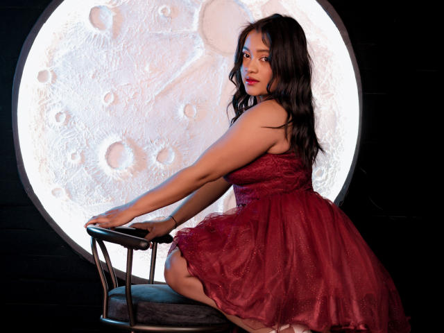 Sarita, avec ses cheveux noirs et ondulés, sa robe en dentelle rouge et son rouge à lèvres rouge, assise sur une chaise, les mains posées sur les accoudoirs, regardant l'appareil photo, avec un cercle évoquant la lune derrière elle..