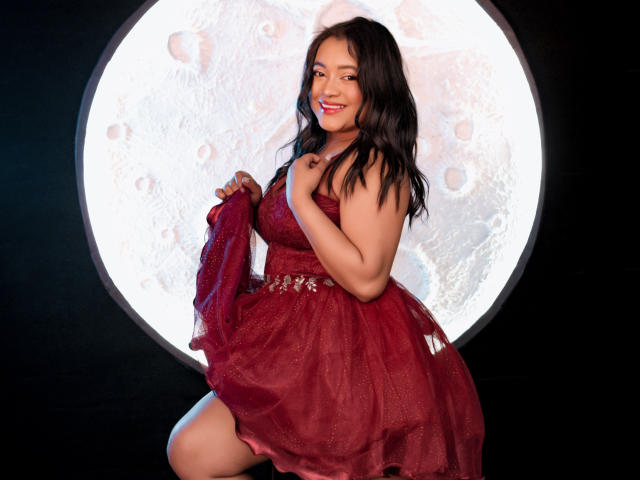 Sarita, carina con un vestito rosso, lo tiene sollevato per mostrare il seno, con un sorriso dolce, capelli neri e uno sfondo lunare in una luce circolare..
