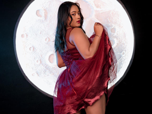 Sarita, une jeune femme aux cheveux noirs et ondulés, portant une robe rouge scintillante, tenant un tissu, regardant par-dessus son épaule, montrant ses formes généreuses, debout devant un grand cercle éclairé par la lune..