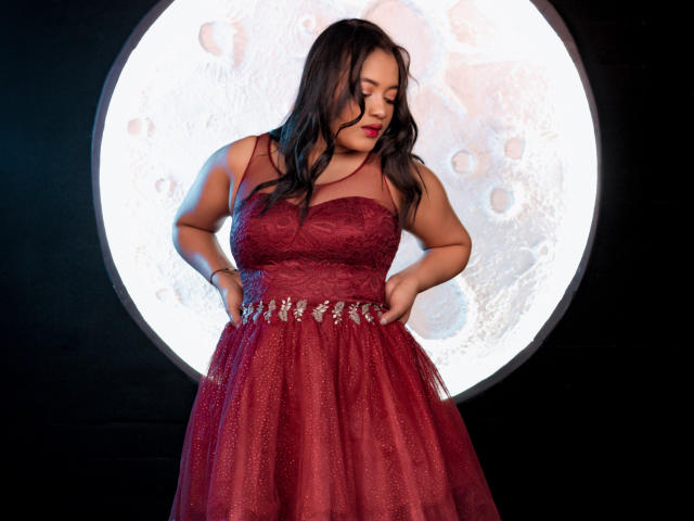 SaritaCute with red lace dress, hands on hips, standing in front of large moon-like light, wavy black hair, red lipstick, and sparkling belt, looking down.