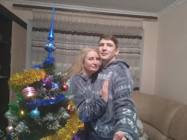 A photograph of JenAndPeter standing in a living room. JenAndPeter, both with fair skin and blonde hair, wear matching grey fleece hoodies. JenAndPeter are positioned in front of a decorated Christmas tree with colorful ornaments and blue tinsel. JenAndPeter's right arms are around each other, with JenAndPeter's left hand on Peter's chest. The background features beige walls, a brown curtain, and a beige sofa. JenAndPeter have relaxed, neutral expressions. The image has a homey, casual feel.