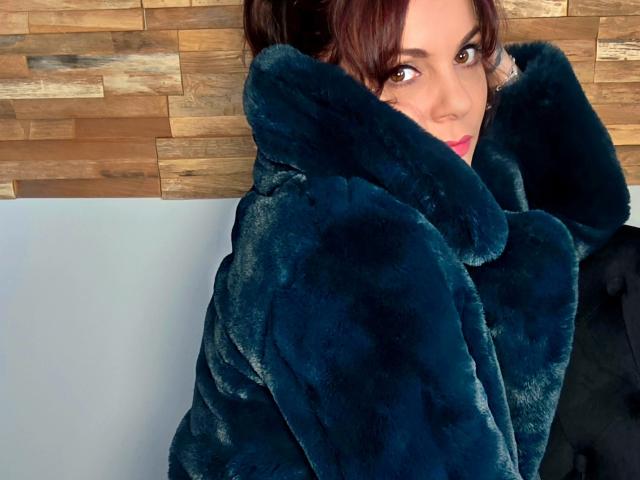 JuneLaFrenchie, with dark brown hair, gazes seductively over her shoulder, wrapped in a thick black fur coat. Wood panel background. She's wearing red lipstick, looking fucking hot.