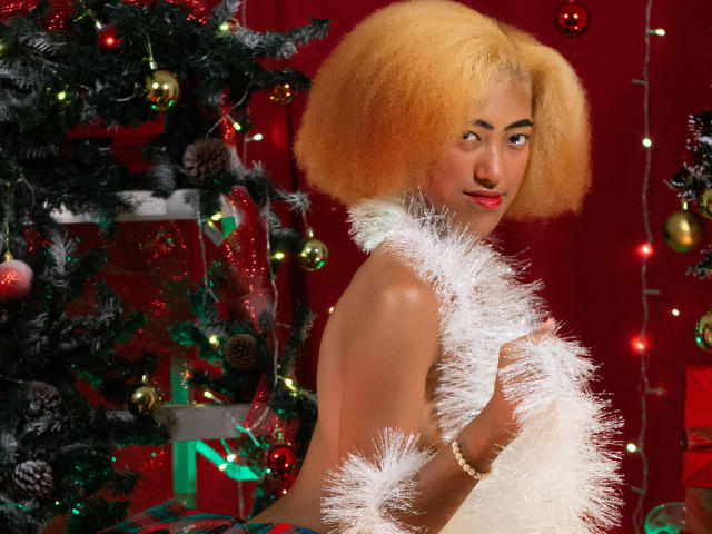 AlishaEbony has a curly, orange-blonde afro and red lipstick. She's topless, holding a white fluffy garland. Her right arm is adorned with a gold bracelet. Behind her, a decorated Christmas tree with red, green, and gold ornaments and a red curtain. The tree has a mix of shiny and textured ornaments, and a gift is partially visible. The image is vibrant, with AlishaEbony's confident, slightly playful expression.