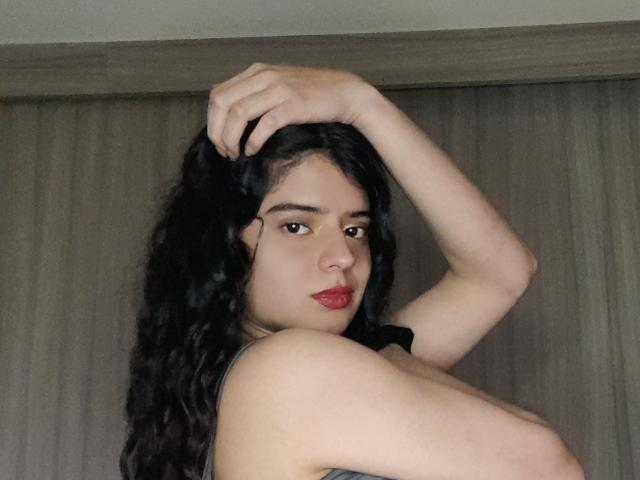 BarbaraTiff with long black curly hair, hand in her hair, looking over shoulder, red lipstick, light skin, bare shoulders, neutral expression, in a photo.