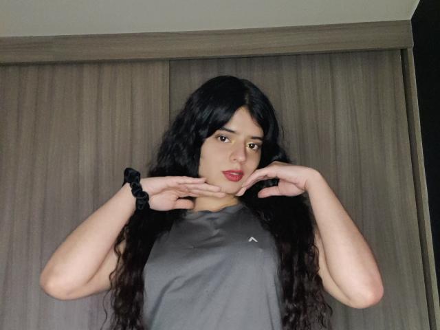 BarbaraTiff with long black hair, gray shirt, and black scrunchie, hands near chin, red lipstick, looking at camera, wooden background, upper body shot, light skin, subtle makeup.