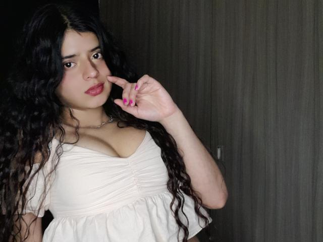 BarbaraTiff with long black curly hair, wearing a white ruched top that shows her big tits, has pink nails, and is touching her lip with her finger.