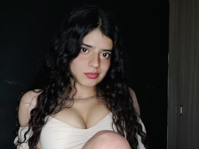 BarbaraTiff with long curly black hair, wearing a white off-shoulder top, showing her big tits, sitting with knees up, looking at camera with slight smile, dark background.
