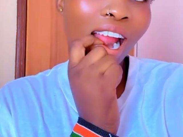 Close-up photo of NattySlim with dark brown skin, showing their face and upper torso. NattySlim has a nose ring in their left nostril, is sticking their right index finger into their mouth, and has white dental braces. They wear a white shirt with a colorful wristband on their right wrist. The light is warm, and the background is blurred. NattySlim's face has a shiny, smooth texture, and their expression is playful.