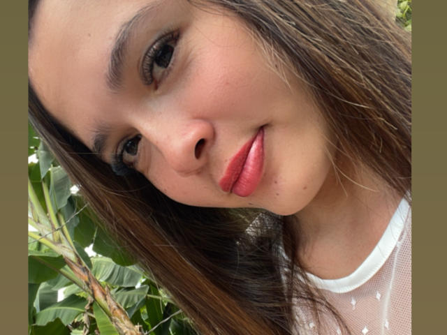 Close-up of Gabrielafoxxy with long brown hair, light brown skin, and pinkish lips. She's looking slightly to the side with a subtle smile. Her dark eyebrows are slightly arched, and she has dark eyelashes. She's wearing a white mesh top with small heart patterns. Green leaves and branches are in the background. The image has a slightly blurred edge and a brown border. The lighting is natural, highlighting her smooth skin and subtle freckles. Her hair is slightly wavy and falls over her right shoulder.