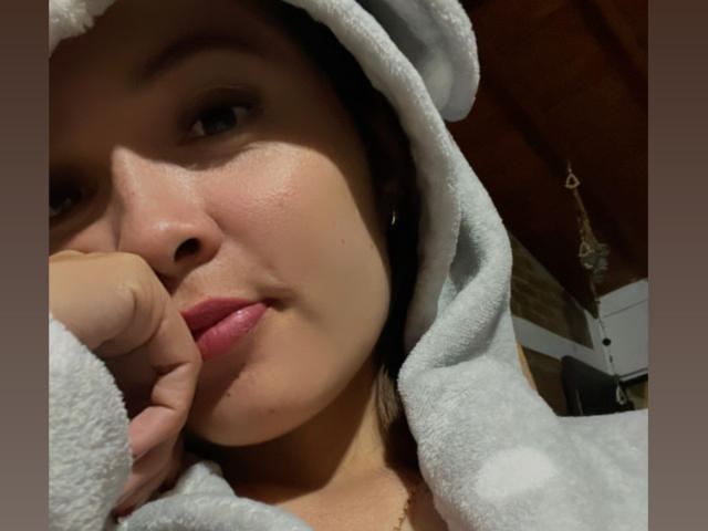 Close-up of Gabrielafoxxy's face, wearing a gray, textured hoodie with a hood. She has light brown skin, subtle freckles, and full, pink lips slightly parted. Her right hand is gently resting on her cheek. She has dark, straight hair partially visible under the hoodie. Her brown eyes are slightly open, and she's looking to the left. The background is blurred, with a wooden ceiling and a hanging decorative item. The image has a soft, warm light. Slight shadow on her cheek adds depth.