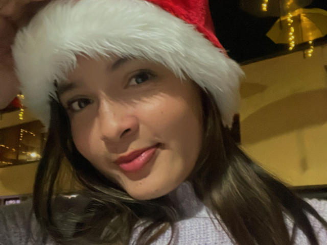 Gabrielafoxxy with a red Santa hat, brown hair, and pink lips, wearing a white-turtleneck, smiling softly. Her tits are not visible. She's cute and sexy.