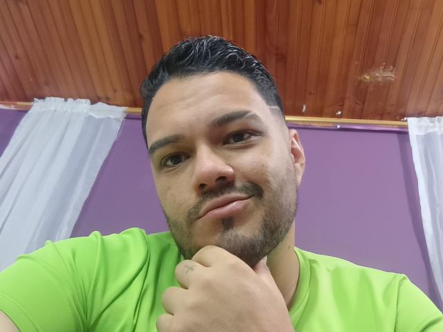 StevenHills, a chubby dude with dark hair and a beard, wearing a green shirt, smiling slightly, touching his chin with his right hand, looking at the camera.