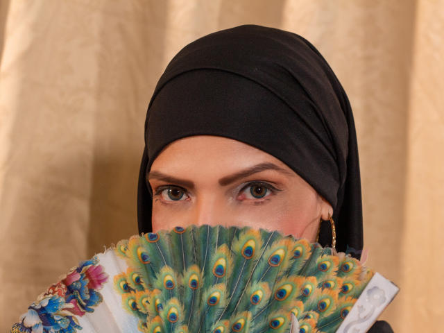 KatteRouse com um leque de pavão cobrindo os seios, usando um hijab preto, olhos verdes, brincos de argola dourados, cortina bege ao fundo..