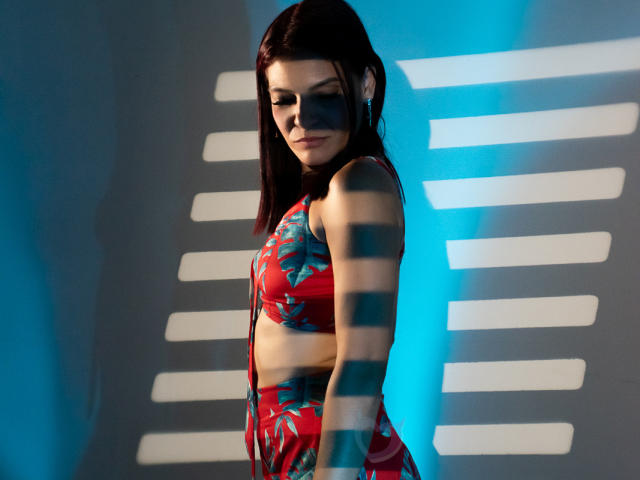 KatteRouse in einem roten, blumigen Bikini, mit blauen Akzentstreifen, im Seitenprofil, nach unten blickend, schlank, helle Haut, dunkelbraunes Haar, kleine Brüste, ein leichtes Lächeln, stehend, blauer Hintergrund..