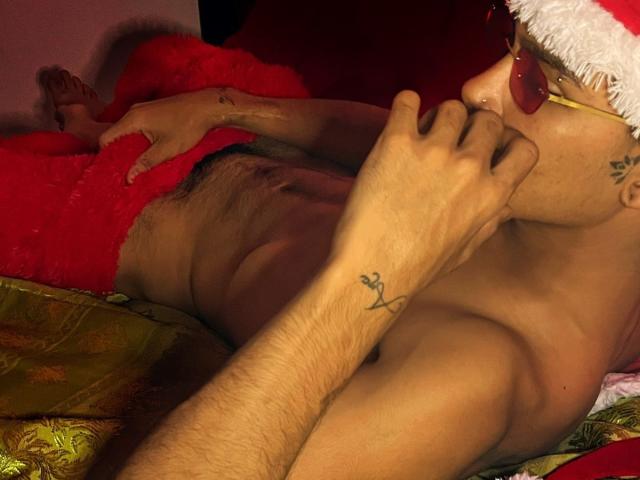 XKamadeva with a Santa hat, red nose, tattoos, and a flaccid penis, lying on a bed, hand near mouth, covering red fuzzy blanket. Male, naked, brown skin.
