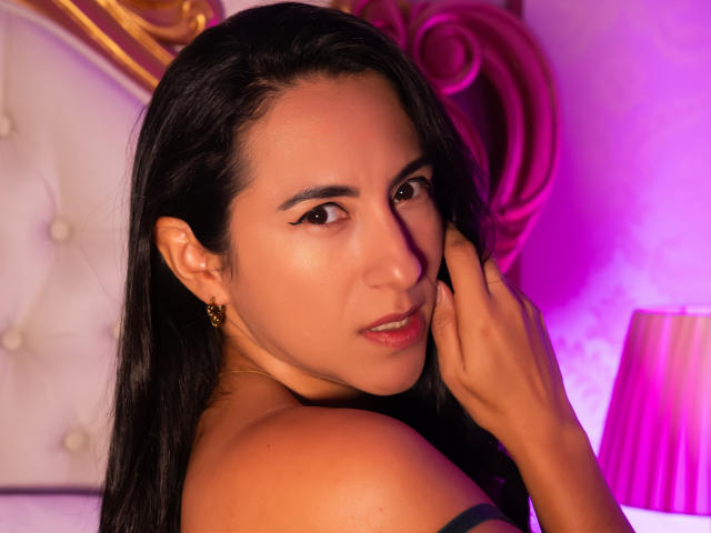 JadeFoster with long black hair, tan skin, and gold earrings, looking over her shoulder, touching her cheek with her hand, showing her shoulder and subtle makeup.