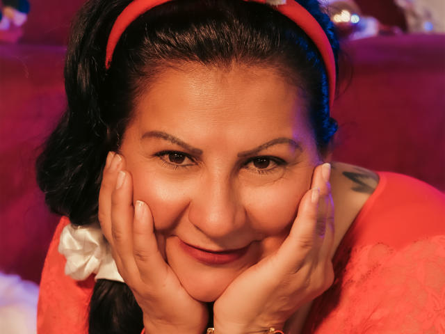 MissLadyy with dark hair in a red headband, hands on her face, smiling, wearing a pink fuzzy top, has a tattoo on her right shoulder, brown eyes.