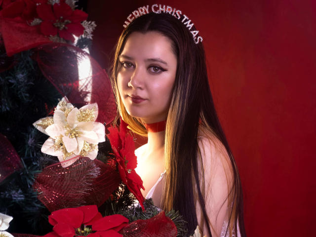 AlayaHot with long brown hair, wearing a red choker and headband saying "Merry Christmas", looking at a Christmas tree with white poinsettia and red ornaments.