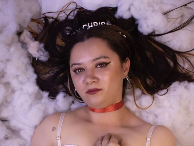AlayaHot with dark hair, red choker, and hoop earrings, lying on white fluff, wearing a tiara, and showing off her tits with a small anchor tattoo on her right.