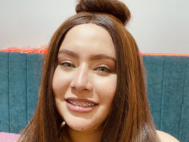 HonneyMil with brown hair in a top knot, wearing metal braces, smiling, green eyes, light skin, blue textured wall in background.