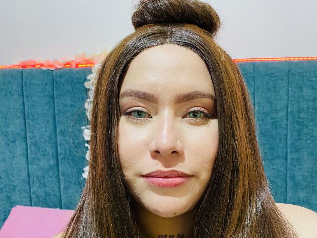 HonneyMil has green eyes, straight brown hair in a bun, and pink lips. She's staring at the camera with a slight smile, her ass is not visible.