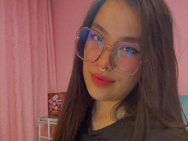 AbbiieMonroe, avec de longs cheveux bruns, du rouge à lèvres, un anneau au nez et de grandes lunettes transparentes, souriant légèrement, portant une chemise noire, avec des rideaux roses en arrière-plan..