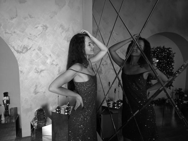 KaraCriegs stands in a dimly lit room, wearing a glittery, strapless dress. She's touching her dark, wavy hair with her right hand, while her left hand rests on her hip. Her reflection in a large, diamond-patterned mirror shows her from the side. The wall behind her has a rough, textured finish. A small, lit Christmas tree and decorated presents are in the background. The black and white photo emphasizes the contrast between her dress and the dark background.