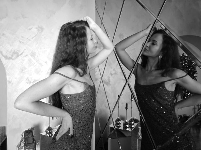 Black and white photo of KaraCriegs standing sideways to a mirror with diamond-shaped reflective panels. She has long, wavy hair and wears a sparkly, sleeveless dress. Her right hand touches her hair, while her left hand rests on her hip. The reflection shows her face and upper body, slightly turned towards the mirror. The room has textured walls and a small, ornate lamp on a table in the background. The image uses high contrast and sharp details to emphasize the texture of the dress and the reflective panels.