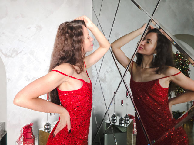 KaraCriegs stands in front of a mirrored wall, wearing a red sequined dress with thin straps. She has long, wavy brown hair and is looking at her reflection with one hand on her head and the other on her hip. The mirror reflects her front and side views. Her skin is fair, and she has a slender build. The room has a Christmas tree in the background. The image has a watermark in the bottom right corner. The lighting is bright, emphasizing the sequins.