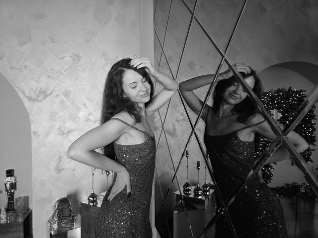 Black and white photo of KaraCriegs. She stands in a room, wearing a sparkly, sequined dress, with one hand in her hair and the other on her hip. Her reflection is in a diamond-patterned mirror. She's smiling, eyes closed, looking relaxed. Bottles are on a shelf to the right. Her long, dark hair is slightly wavy. The textured wall is in the background. The mirror's geometric pattern contrasts with the wall's rough texture. The overall composition is balanced and reflective.